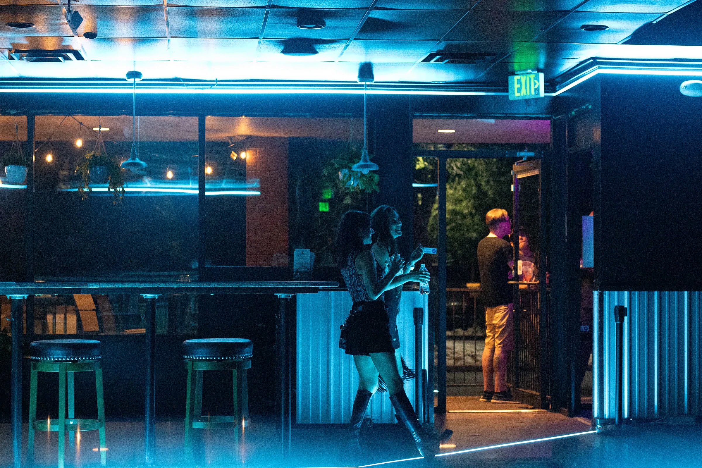 The front bar and entrance at The Midnight Toad at night, lit in teal neon, with the door open to the street.