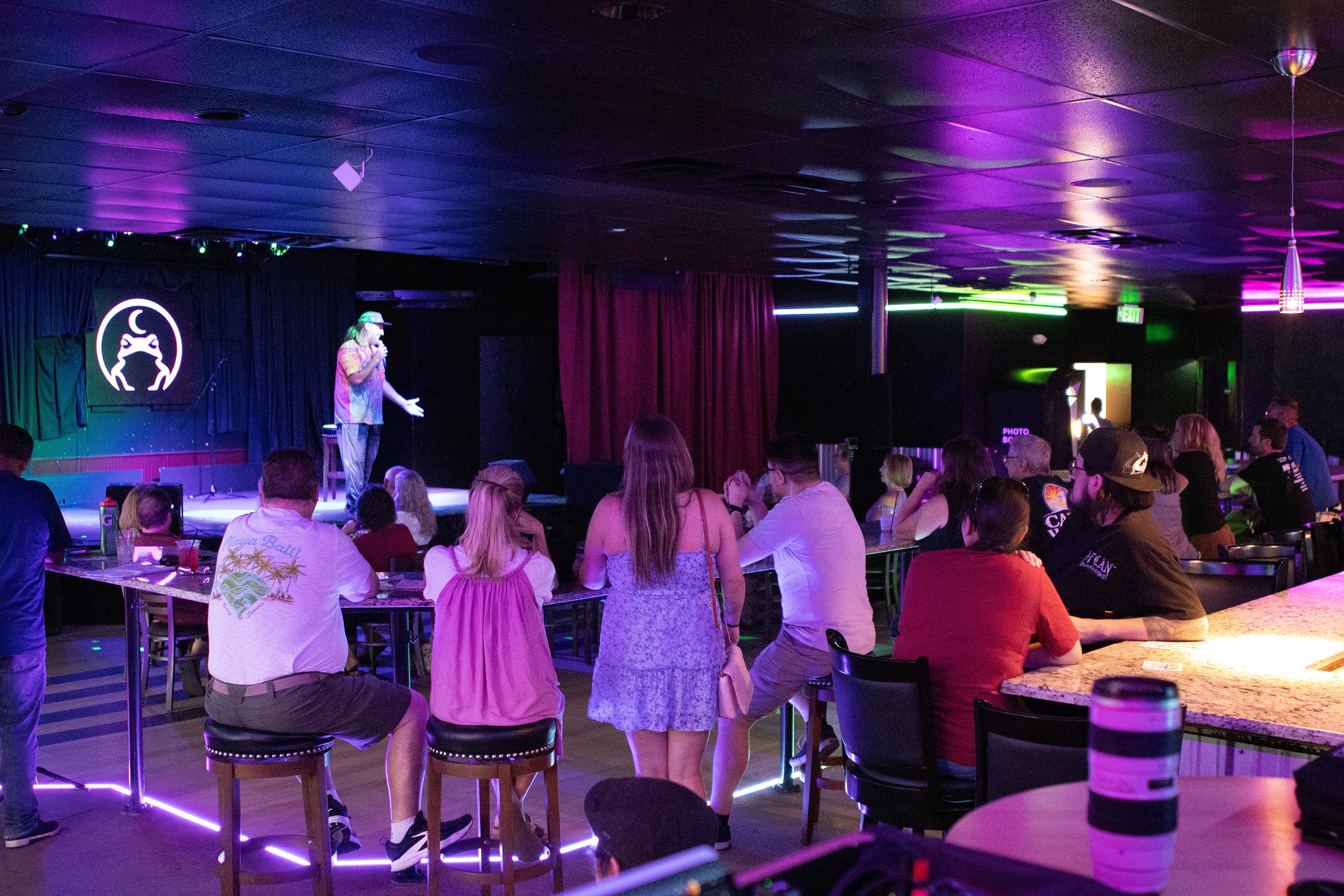 A comic on stage at The Midnight Toad with a full room watching from the bar and the tables, the whole room washed pink and purple.